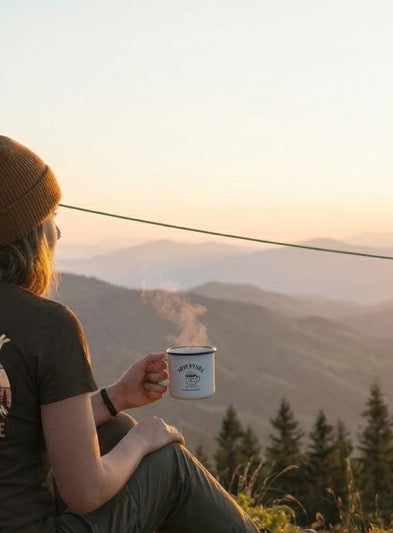 Person sitting outdoors with a coffee cup, mountains in the background, and a t-shirt with nature design.